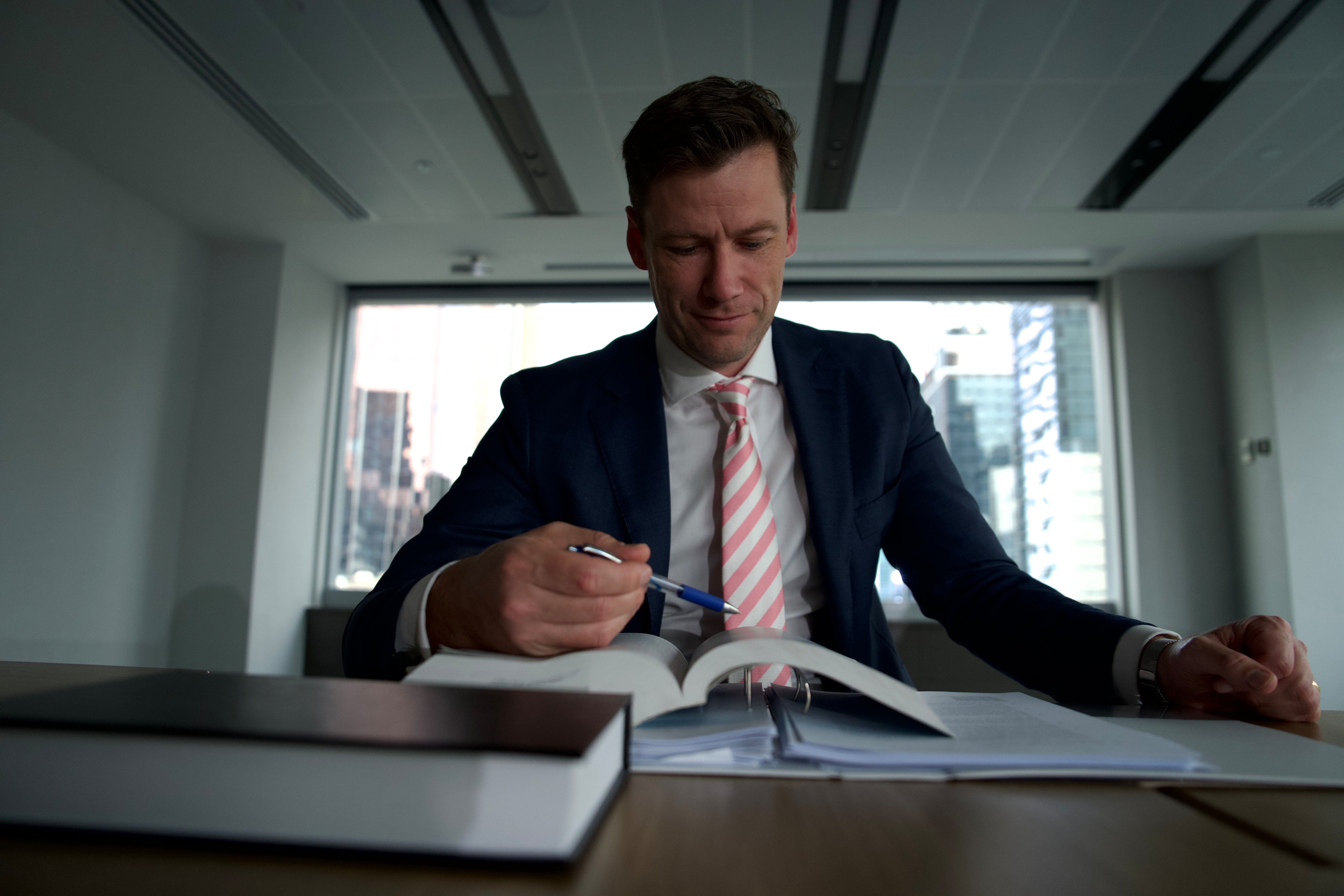 Joel Phibbs sits at a desk with an open book and pen in his hand his back is to a large window