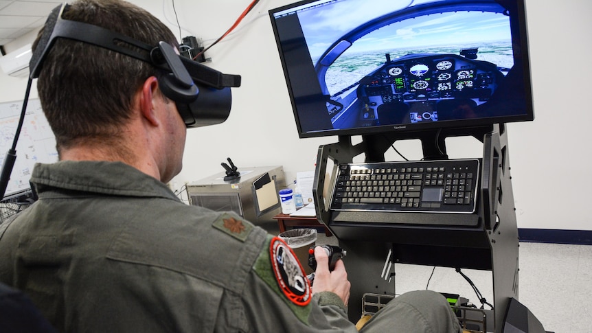 The back of a military trainee wearing virtual reality goggles sitting in a simulator with computer screen