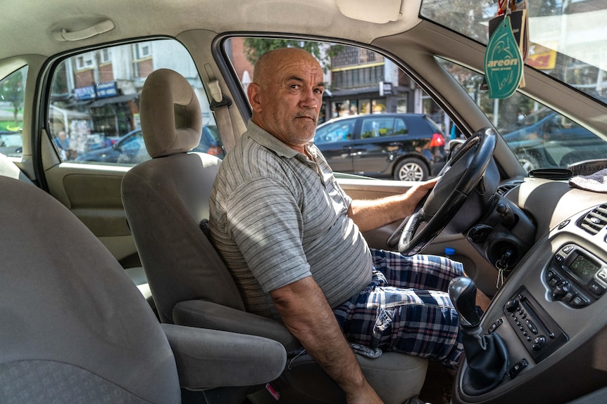 A bald man wearing a striped shirt and chequered pants looks over his shoulder in the drivers seat of a car.