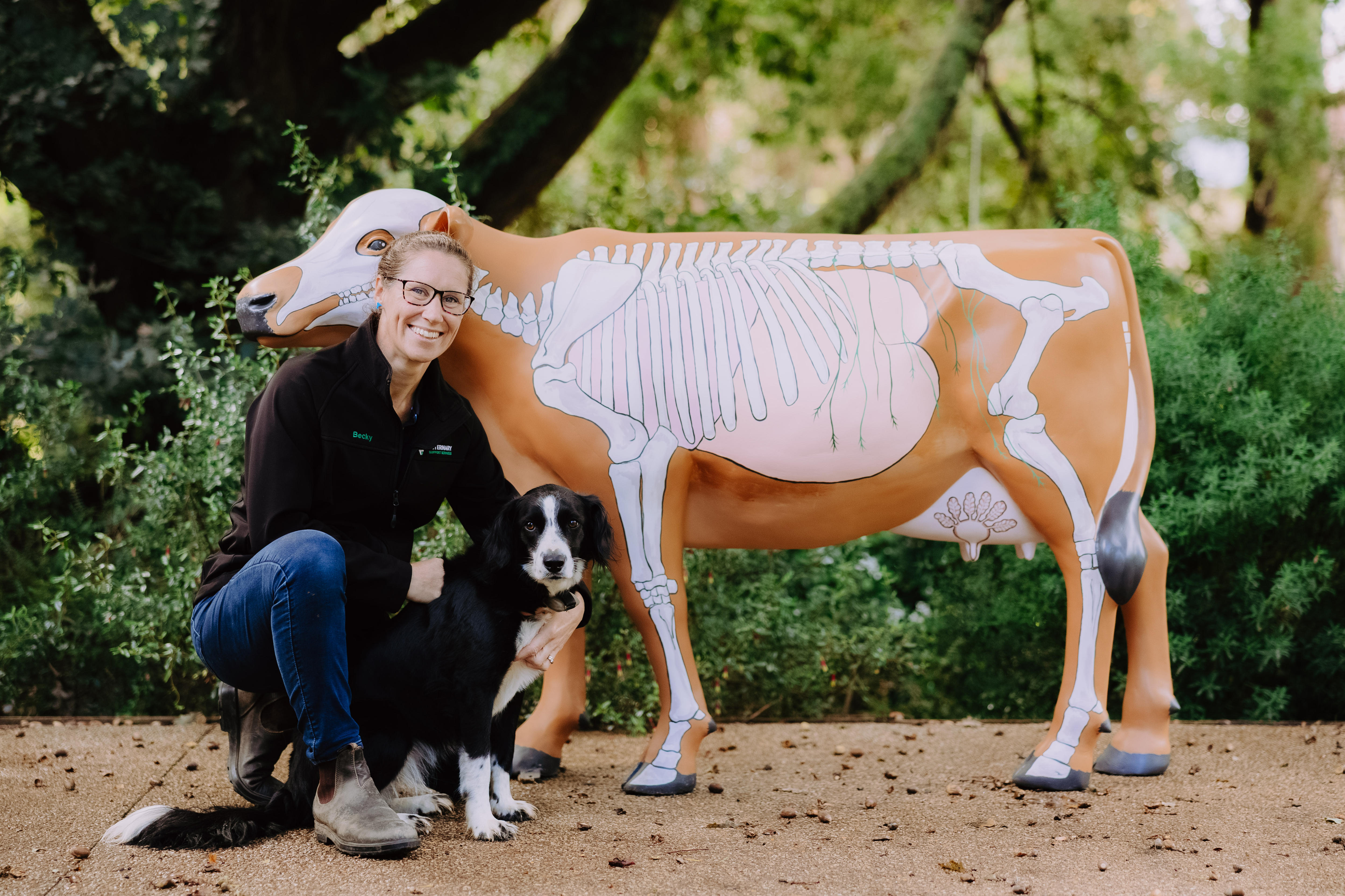 A woman in a black shirt poses next to a fake cow with a dog