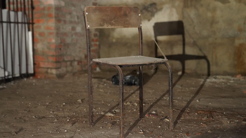 An old chair with metal legs and a wooden seat and back sits on the dirty floor of a basement. A cobweb extends between corners
