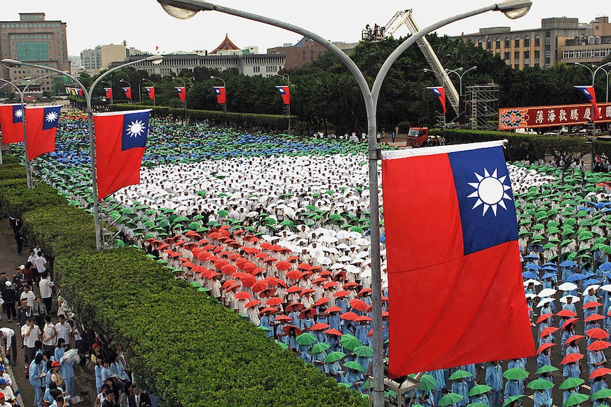 A parade of students in different coloured 'umbrella hats' parade up a wide street where Taiwanese flags hang from street lamps