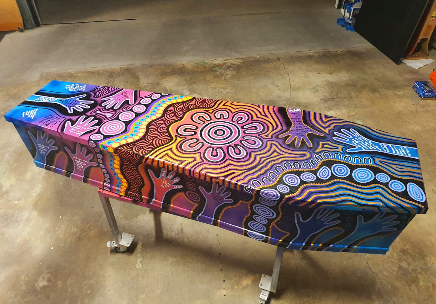 A coffin propped up on metal legs painted in bright fluro patterns and traditional Aboriginal dot painting.