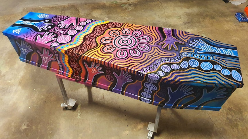 A coffin propped up on metal legs painted in bright fluro patterns and traditional Aboriginal dot painting.