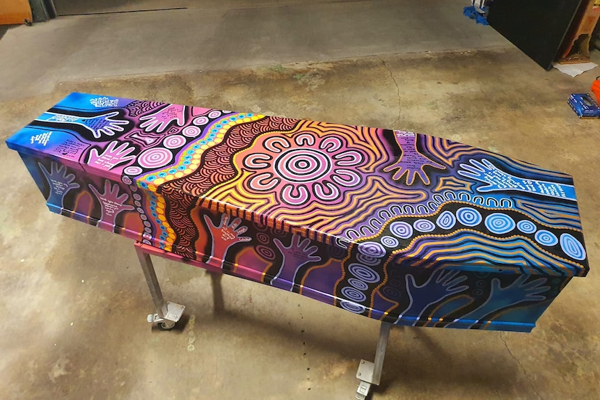 A coffin propped up on metal legs painted in bright fluro patterns and traditional Aboriginal dot painting.