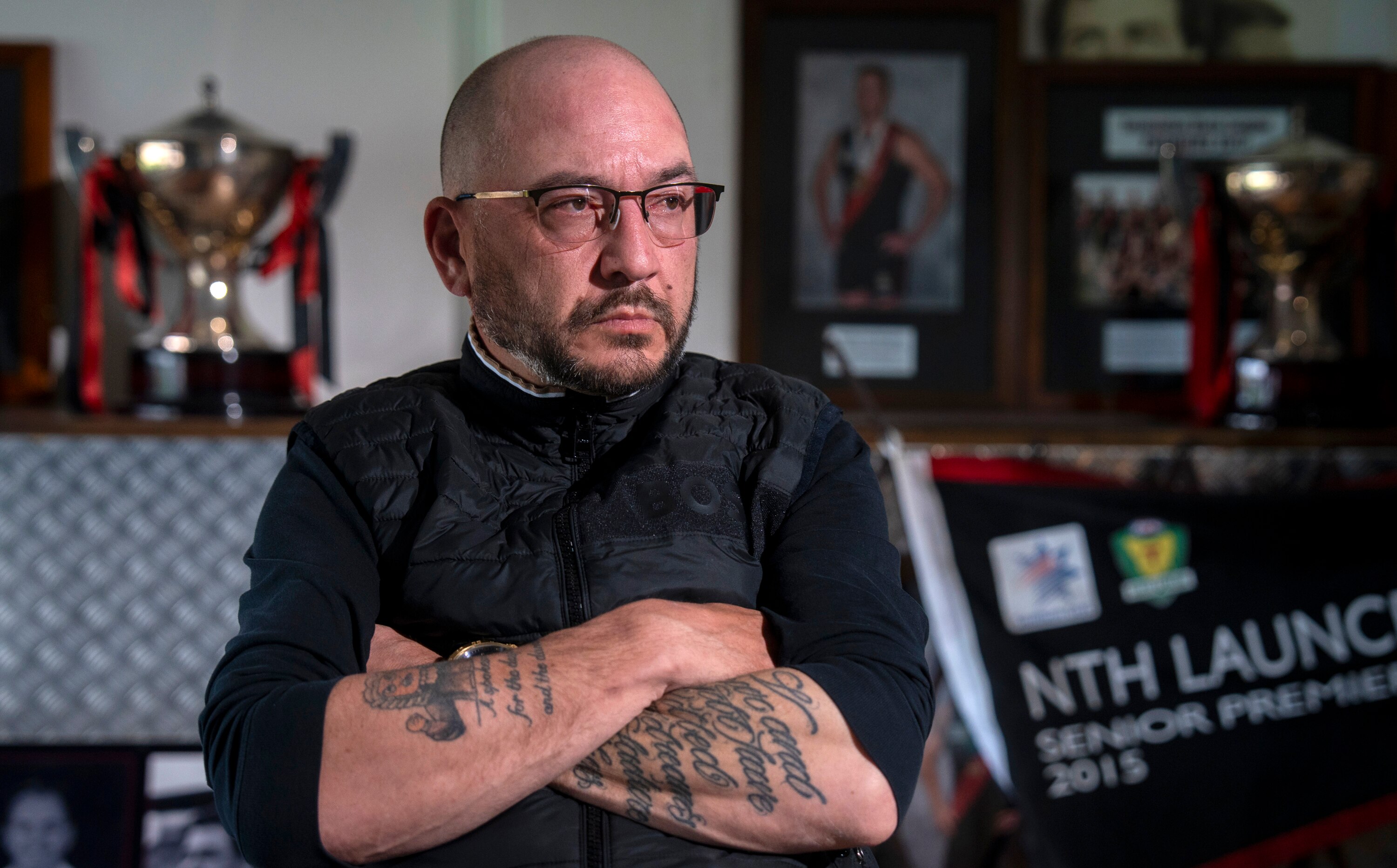Un hombre con gafas y tatuajes en las mangas está sentado con los brazos cruzados y trofeos y premios al fondo.