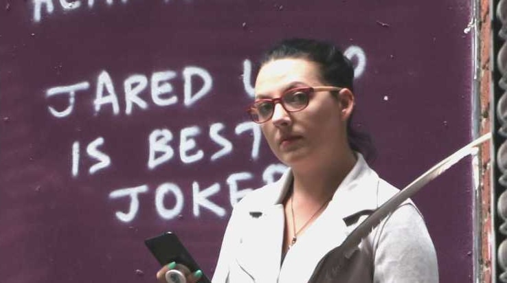 A picture of a woman holding a phone looking at the camera. Graffiti art on the wall behind shows a clown.