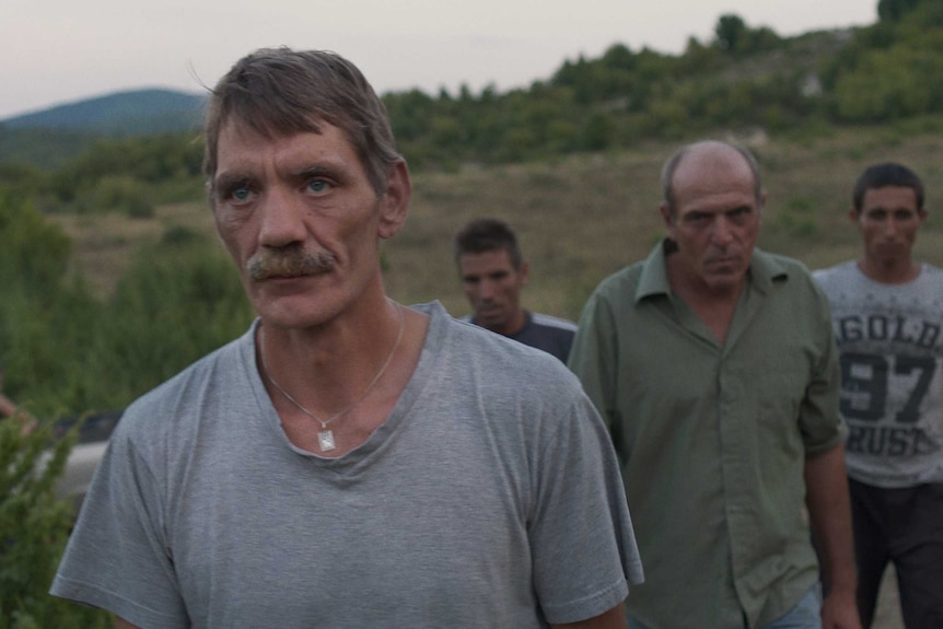 A group of older men walk in the shadowy country-side, in low light.