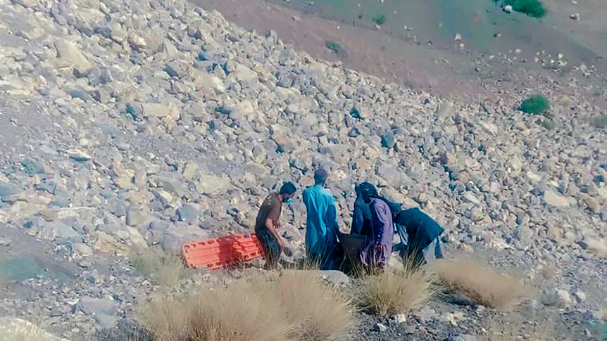 Five men with a stretcher stand on a rocky, arid mountain slope