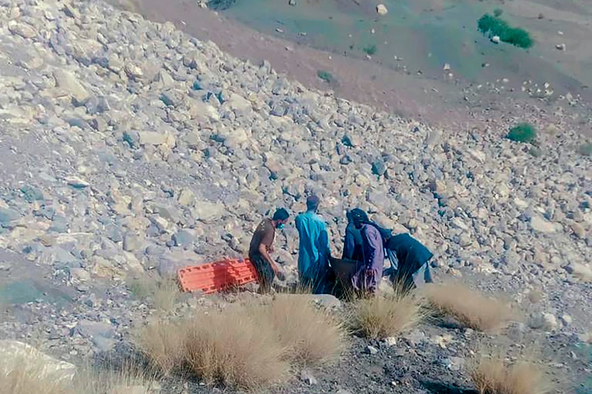 Five men with a stretcher stand on a rocky, arid mountain slope