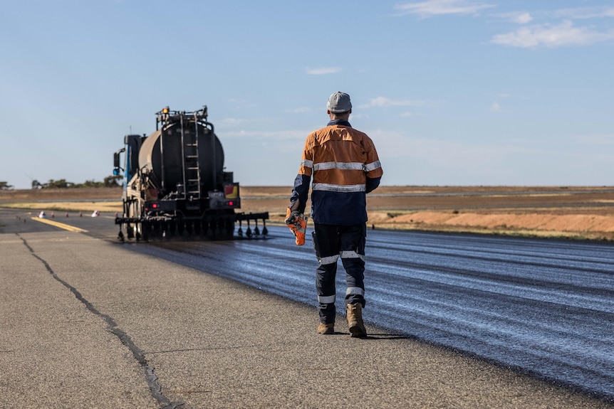 A worker in high-vis walks behind a track resurfacing a bitumen runway at an airport.  