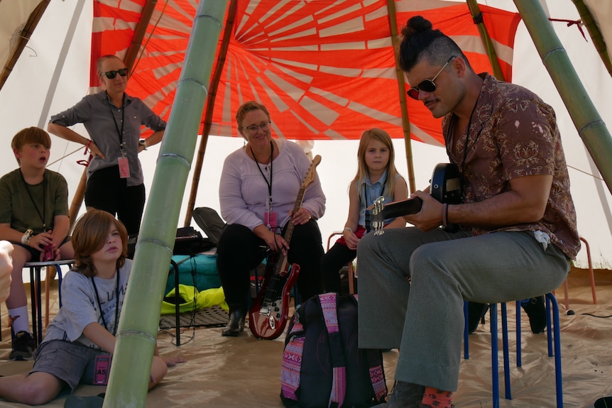 A man with a guitar in a tent with primary school students holding instruments