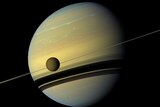 The planet Saturn appears against the jet black of space, with its rings side-on and a moon in the foreground.