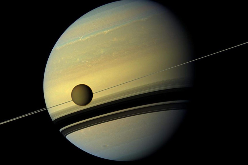 The planet Saturn appears against the jet black of space, with its rings side-on and a moon in the foreground.