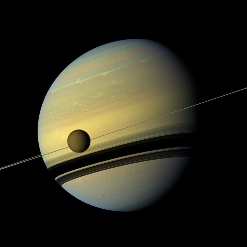 The planet Saturn appears against the jet black of space, with its rings side-on and a moon in the foreground.