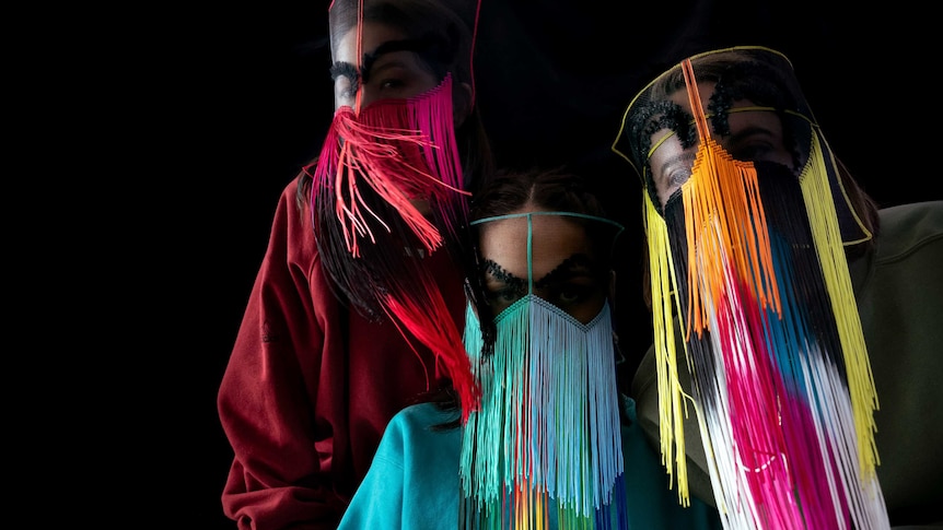 Haiku Hands (three women) wear mesh, full-face coverings with rainbow coloured fringing. Background is black.