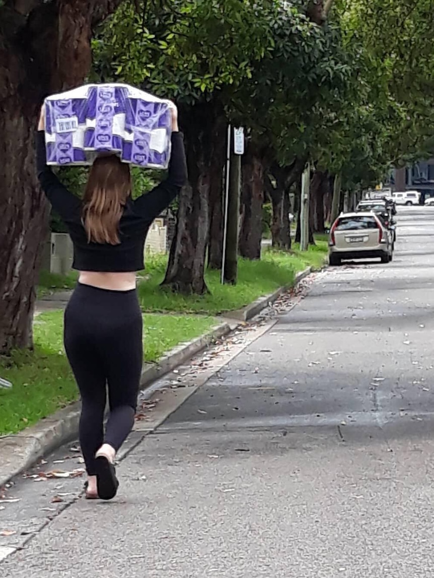 A woman carries a bulk pack of toilet paper above her head as she walks down the street.