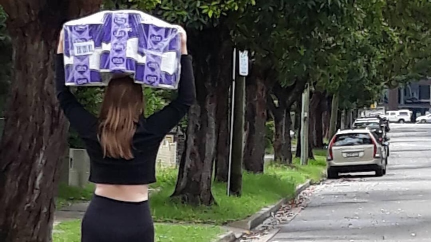 A woman carries a bulk pack of toilet paper above her head as she walks down the street.