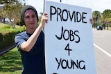 A young man wearing a beanie holds a sign that reads "provide jobs 4 young people".