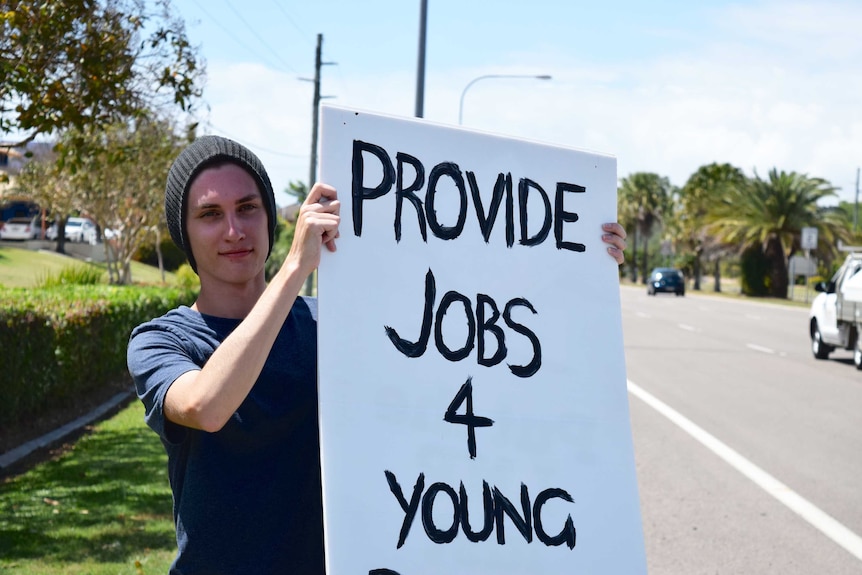 A young man wearing a beanie holds a sign that reads "provide jobs 4 young people".