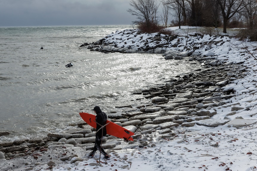 A surfer in a full wetsuit carrying a red board walks through snow to an icy lake shore 
