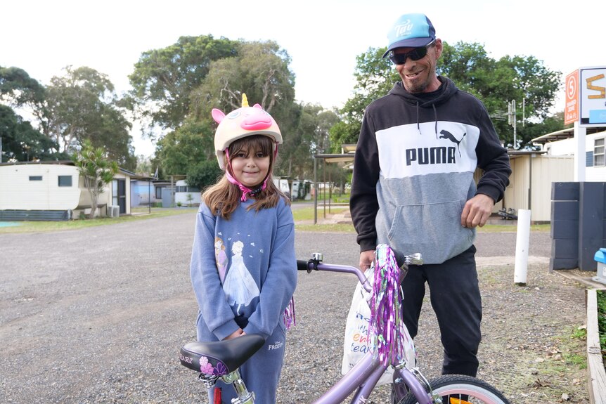 Dad and daughter standing on a road in a caravan park with her new purple and pink bike with tassels and her helment on.