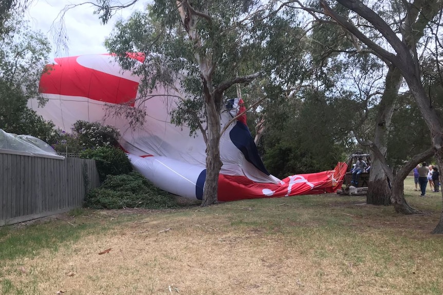 A red and white hot air balloon lies in parkland, partially draped over the fence of a neighbouring house's backyard.
