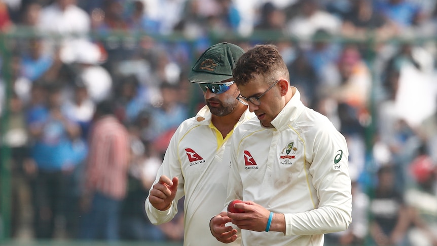 One Australian spin bowler leans in and talks to another spin bowler who is holding the ball walking back to his mark in a Test.