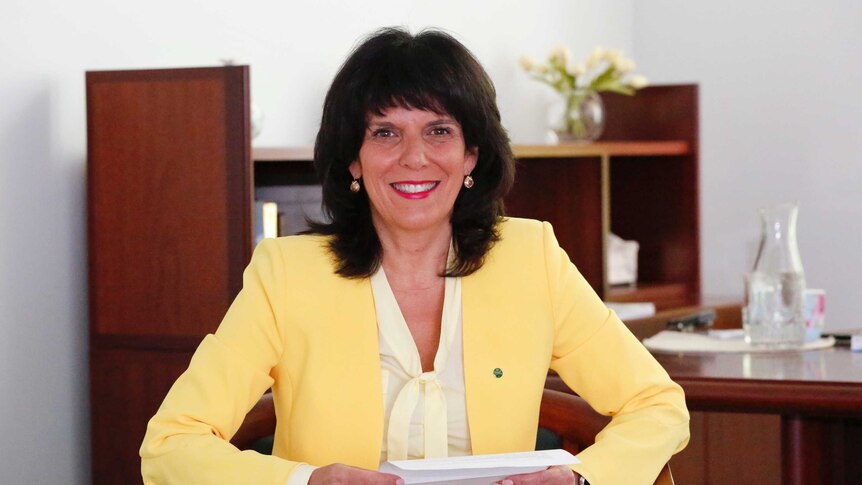 Julia Banks, wearing a lemon yellow jacket, sits in a chair in her office smiling. She is holding a piece of paper in her hands.