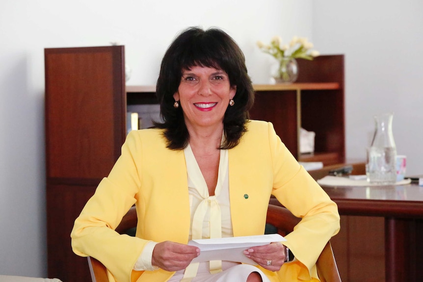 Julia Banks, wearing a lemon yellow jacket, sits in a chair in her office smiling. She is holding a piece of paper in her hands.