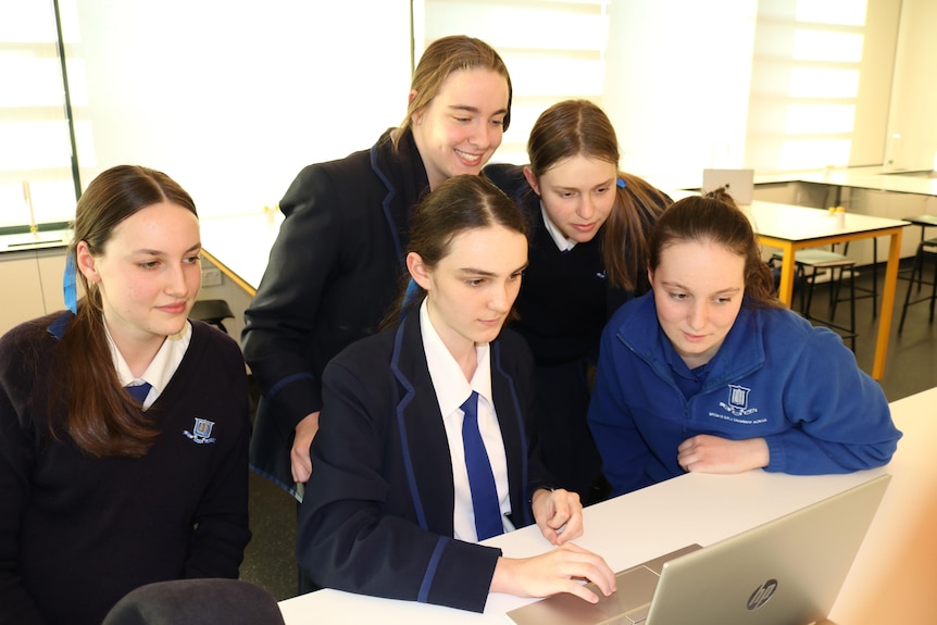 An image of five students huddled around a laptop. 