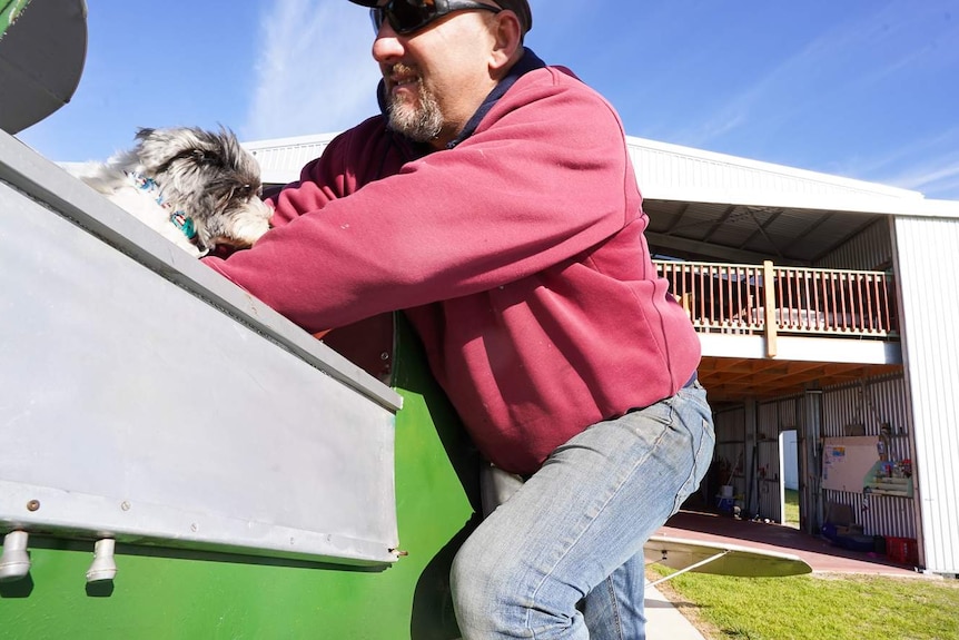 A man in a bear, sunglasses and cap lifts a small dog into the cockpit of a plane while smiling.