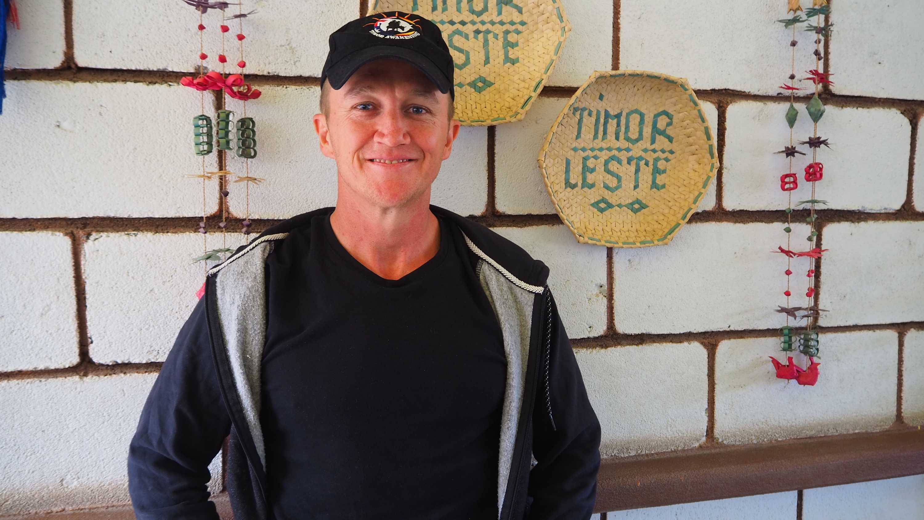 A man is smiling in front a brick wall he is wearing a black hat, shirt and jacket
