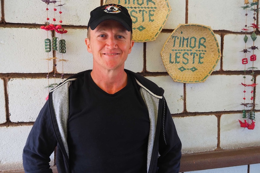 A man is smiling in front a brick wall he is wearing a black hat, shirt and jacket