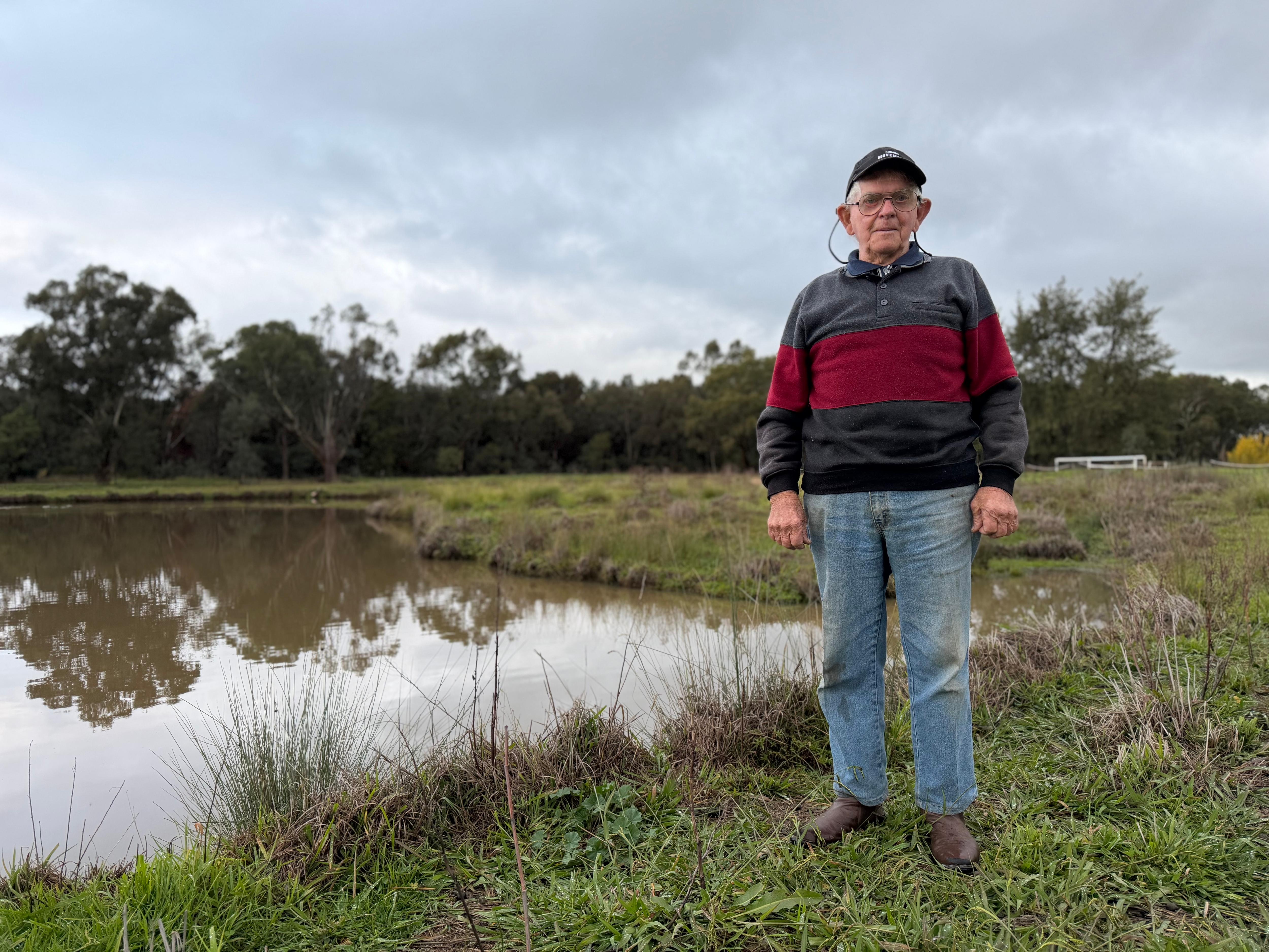 An older man wearing glasses and a cap stands beside a small dam. 