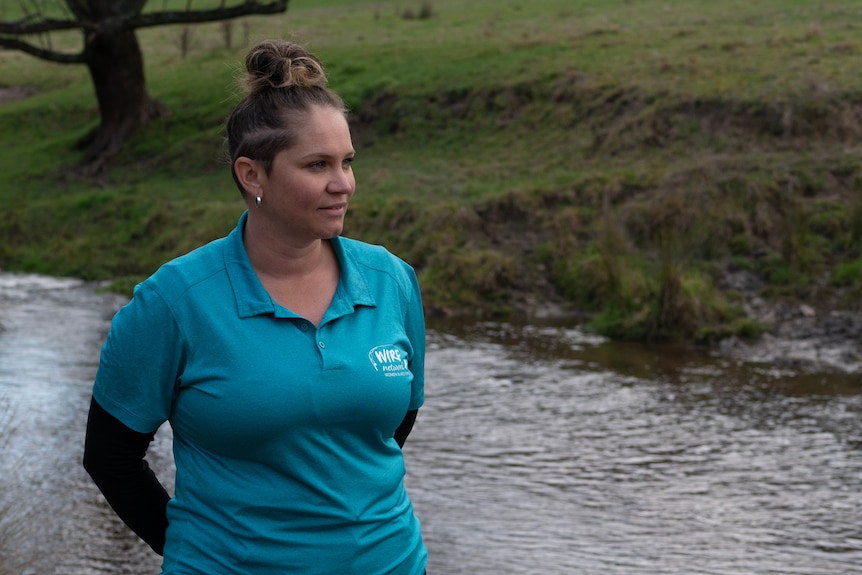 a woman with hair a top pony and undercut, wearing a blue shirt stands next to a creek