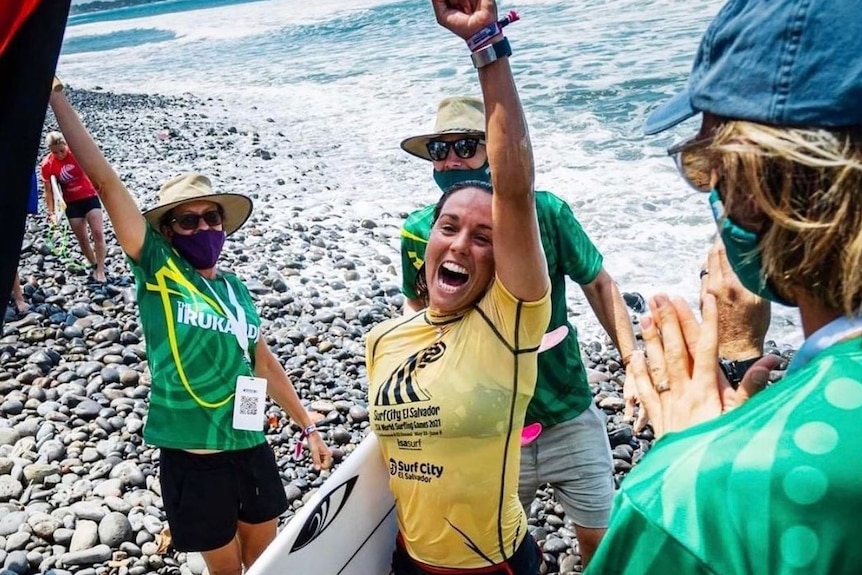 Woman holding a surf board pumps her fist in the air and is cheered on by people around her.