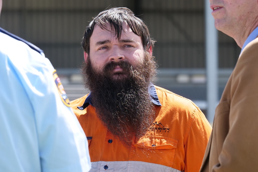 A man wearing a high visibility orange work shirt with a long black beard standing next to two men slightly out of frame