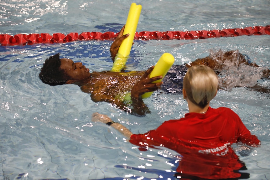 A man in the swimming pool with a yellow pool noodle and an instructor in a red rash vest