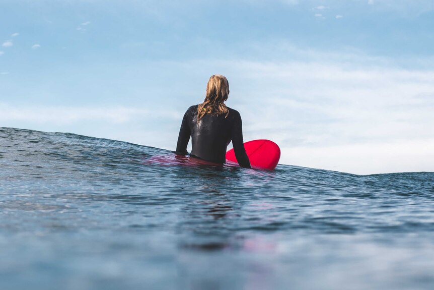 A woman wearing a wetsuit sits up on a red surfboard to depict how to choose your first surfboard.