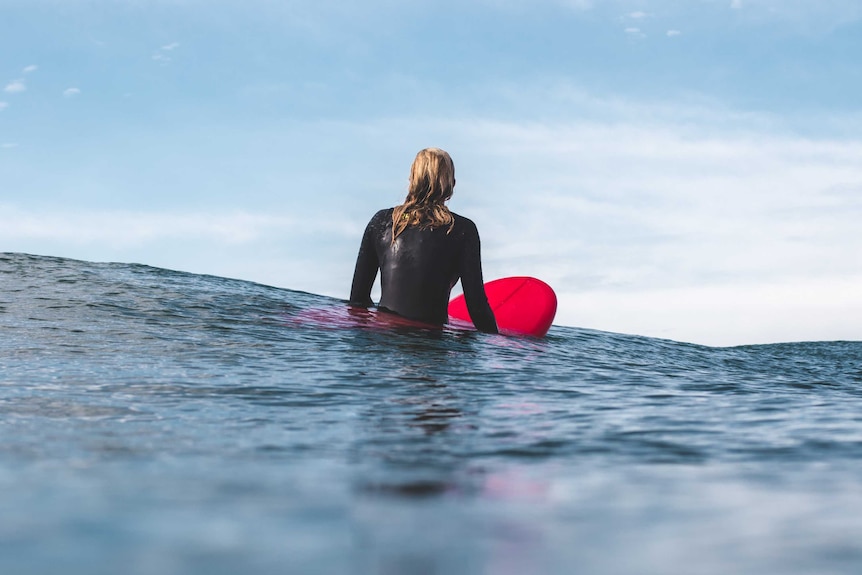 A woman wearing a wetsuit sits up on a red surfboard to depict how to choose your first surfboard.