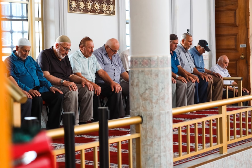 Man sit on a bench in a mosque with their heads lowered. 