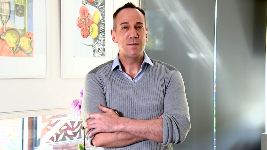 a man with his arms folded looks at the camera while standing in his kitchen