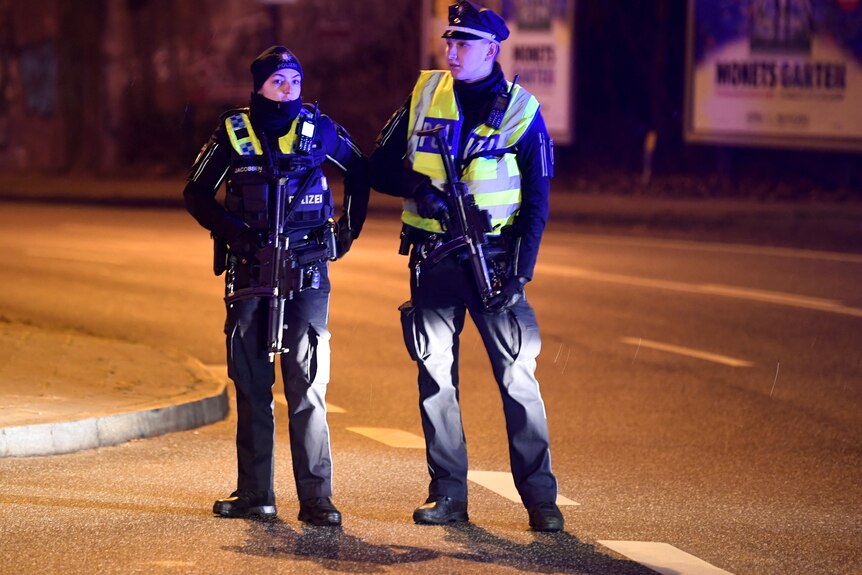 Two officers satnd holding weapons next to a street at night. 