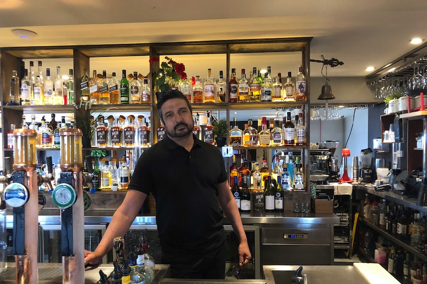 A man leans against a bar with a large array of various alcohol bottles behind him. Two beer taps are in the foreground.