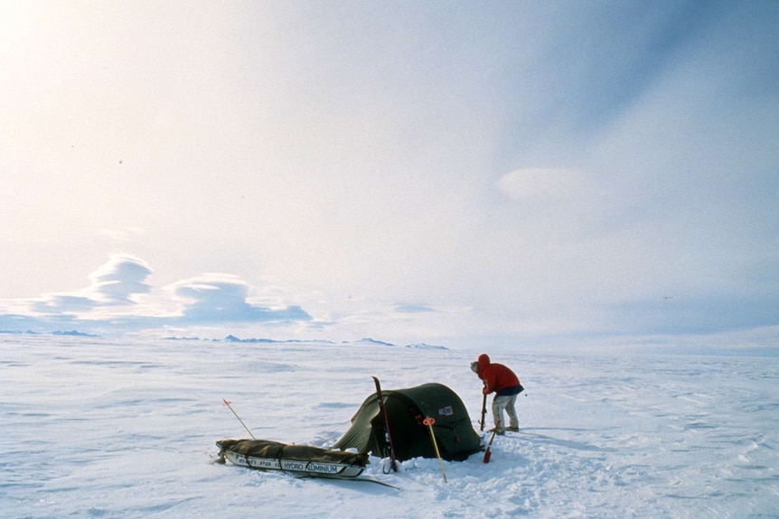 A lone man stands with a small tent and sled in the vast Antarctic landscape.