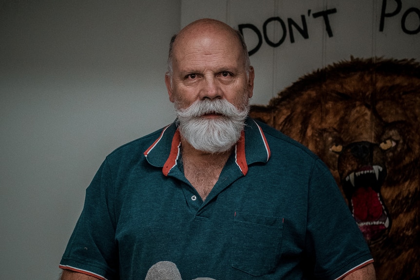 A Balding man with a white moustache and green polo shirt in front of an angry bear mural.