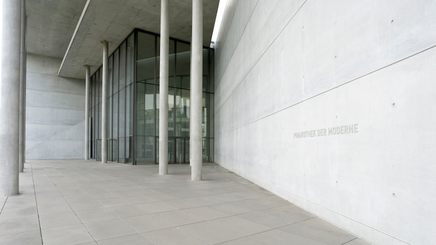 The entrance of a modern concrete building with high glass doors and windows, text on the wall reads "Pinakothek der Moderne"