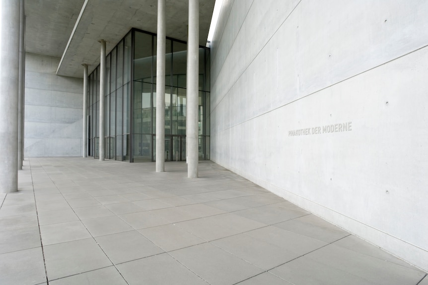The entrance of a modern concrete building with high glass doors and windows, text on the wall reads "Pinakothek der Moderne"
