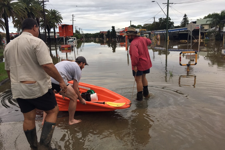 Three man stand around a small orange boat being pushed into floodwaters along a  street lined with shops
